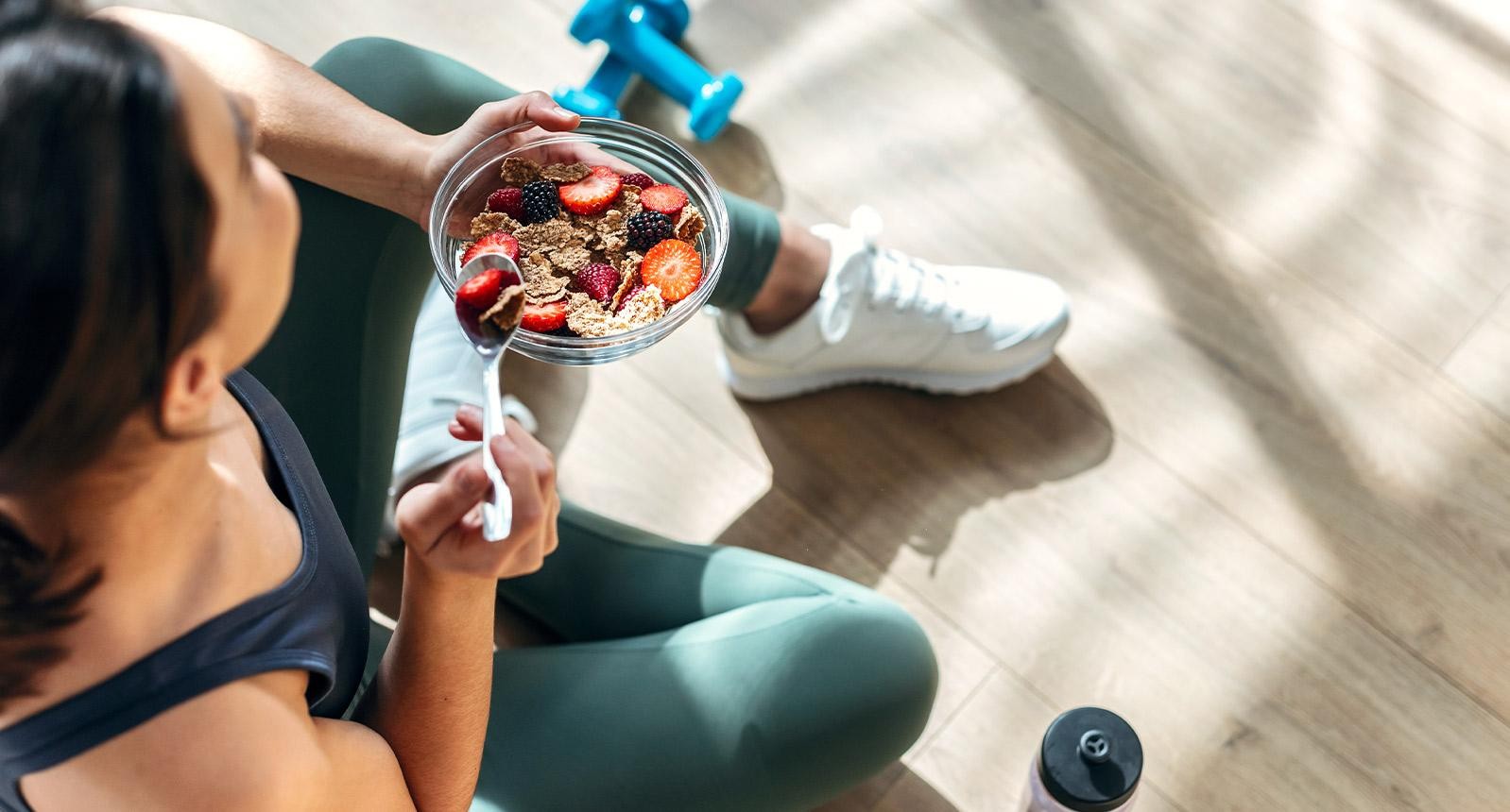 Shot of athletic woman eating a healthy bowl of muesli with fruit sitting on floor in the kitchen at home