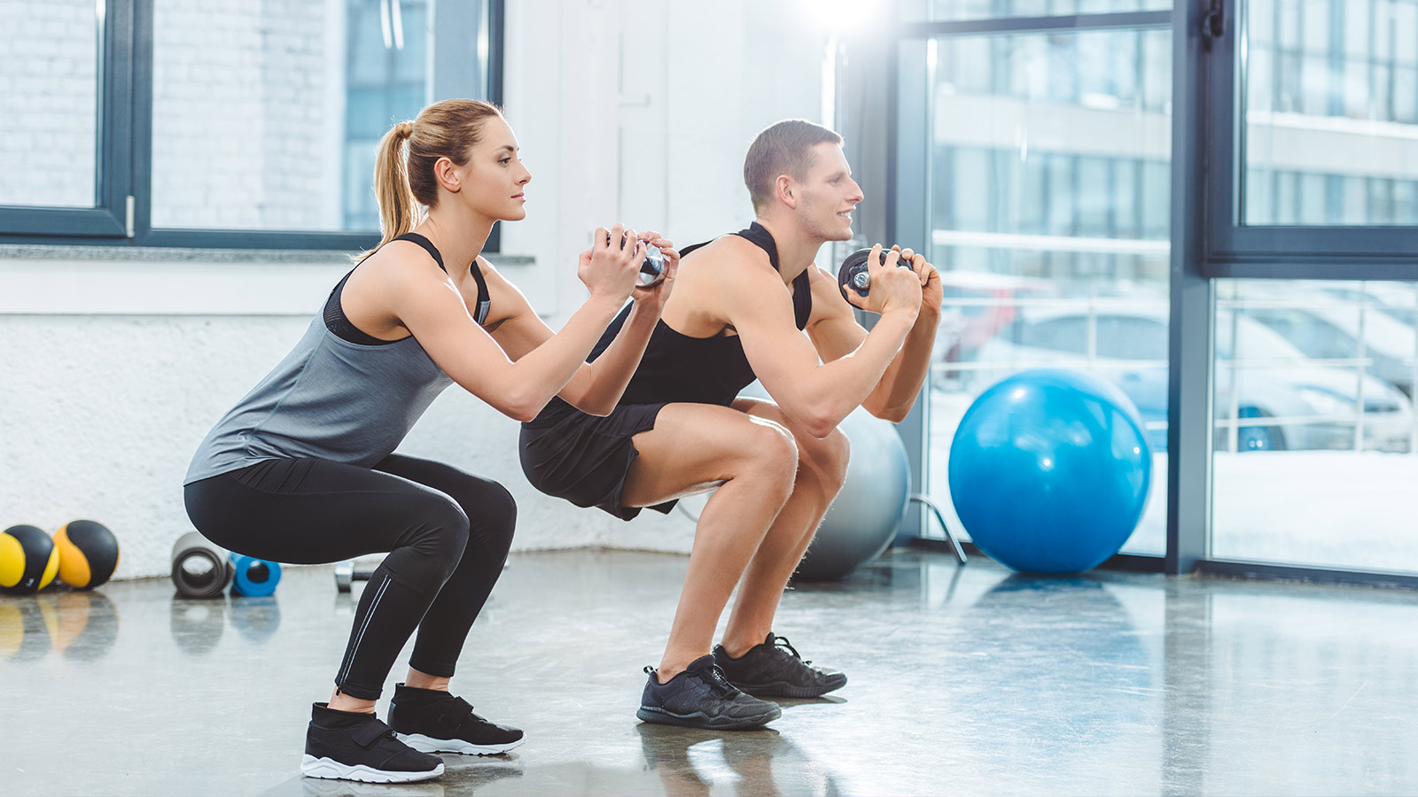 sporty young couple holding dumbbells and doing squat exercise in gym